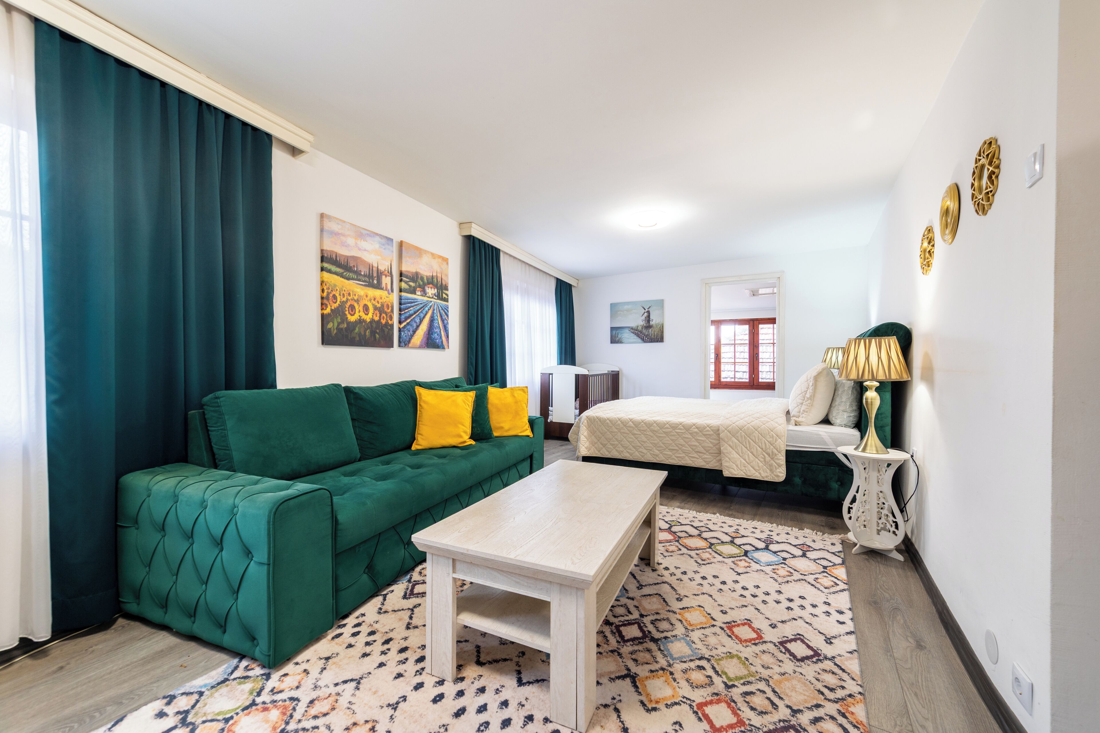 Een kamer met een groene bank, een witte tafel, een bed en een kamerkleed met dessin.