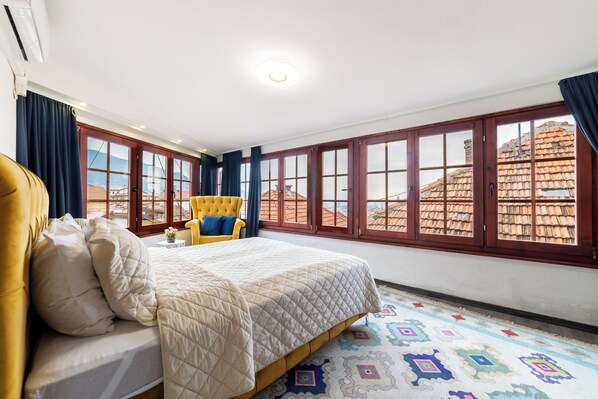 Un dormitorio con cama, una silla, ventanas amplias y vistas a edificios.