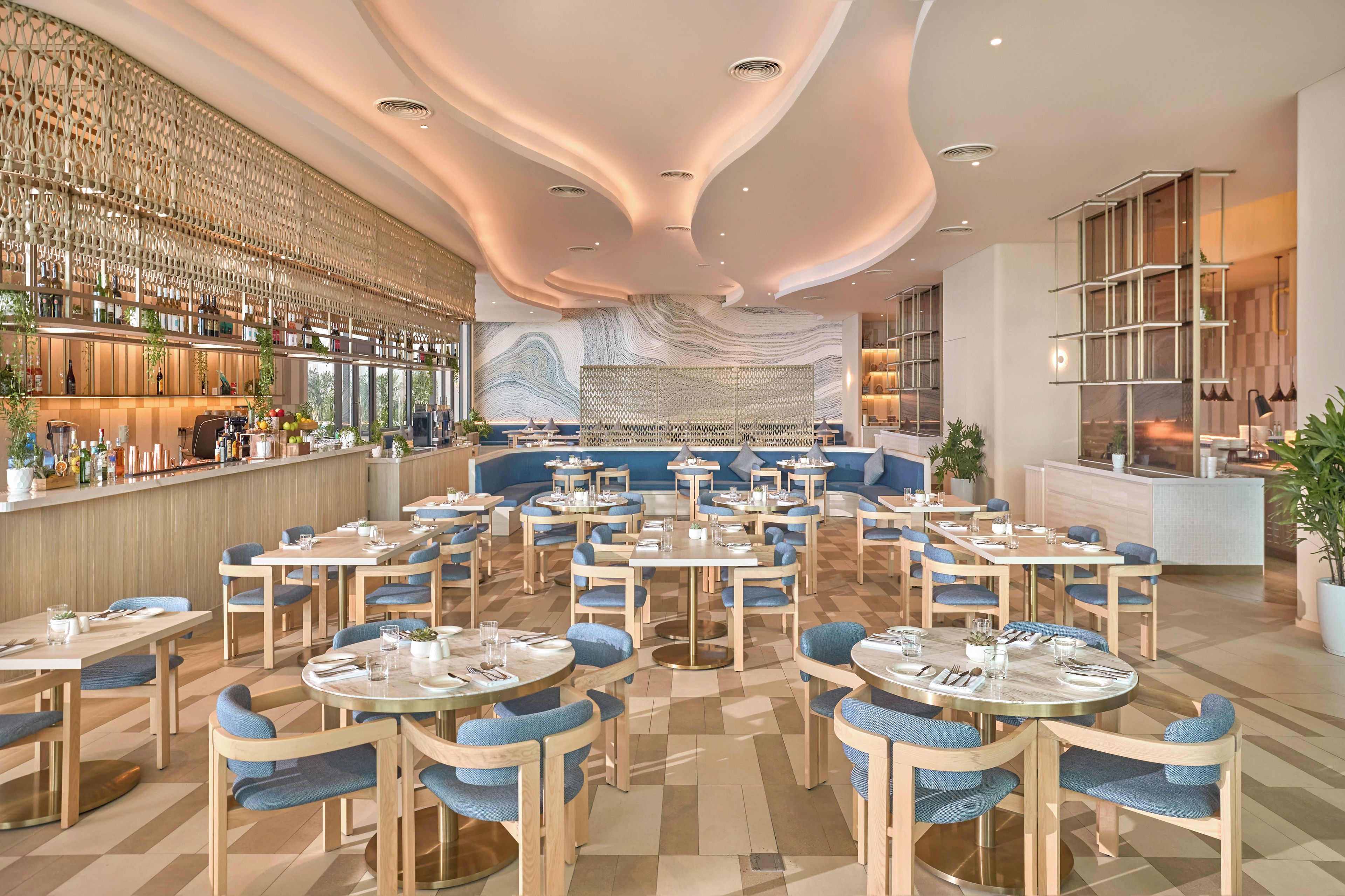 A modern restaurant with round tables, blue upholstered chairs, and a bar area with shelves of bottles.