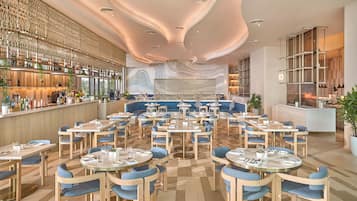A modern restaurant with round tables, blue upholstered chairs, and a bar area with shelves of bottles.