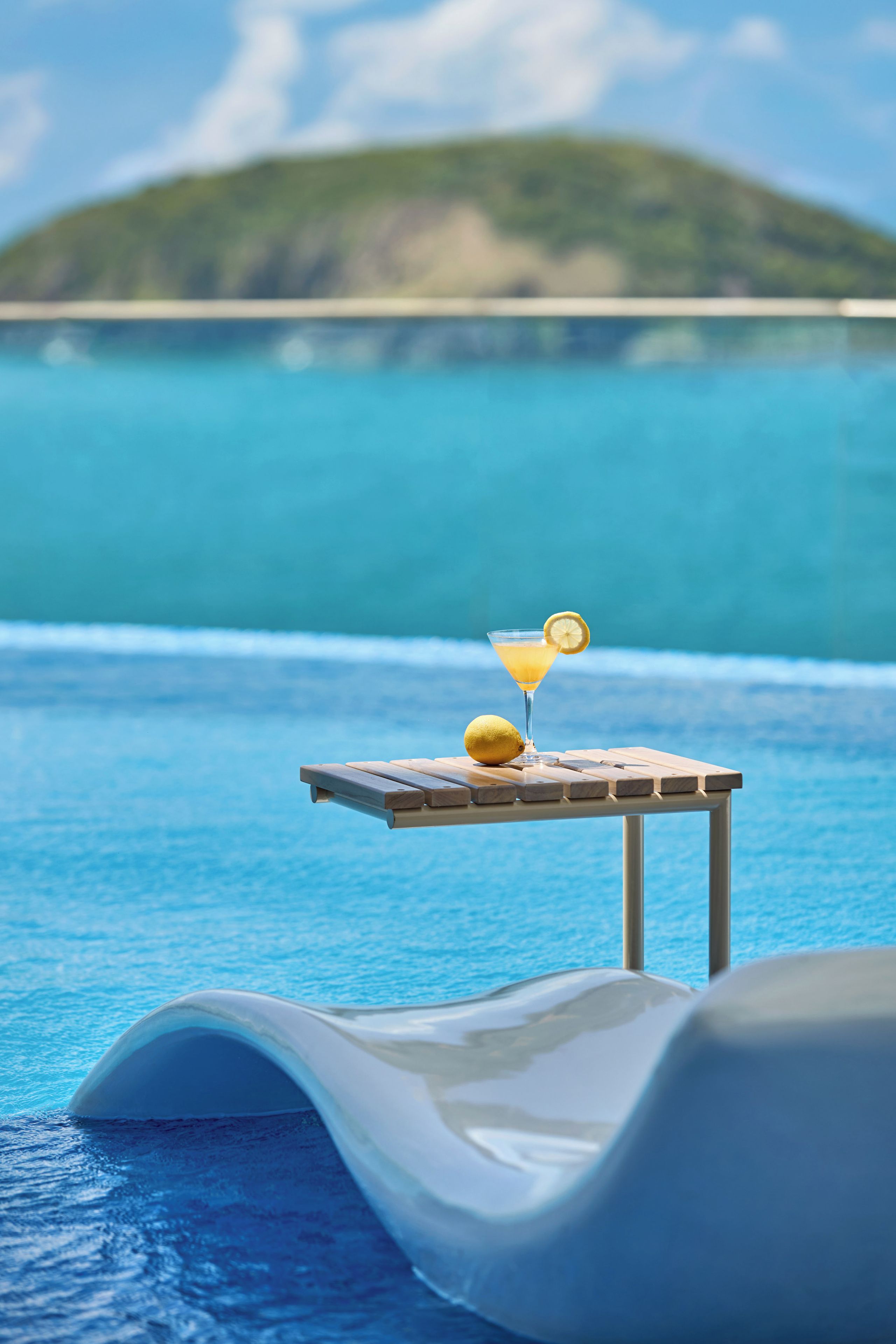 A poolside bar with a glass of cocktail and a lemon slice, overlooking a clear blue pool and a distant island.