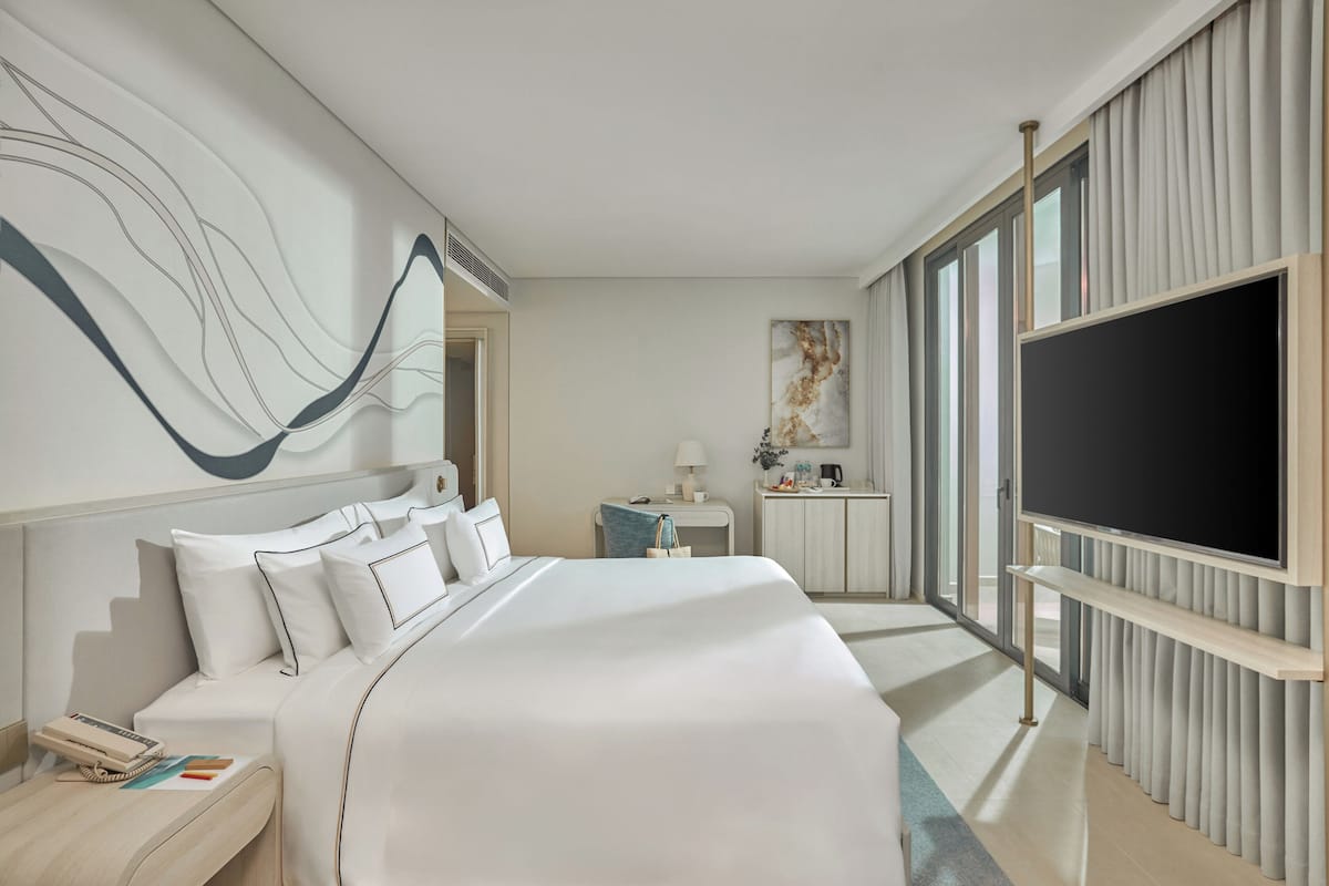 A modern hotel room with a large bed, a flat-screen TV, a desk with a lamp, and a view through large windows.