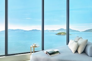 A room with a large window overlooking a body of water and distant mountains. There is a bed with pillows and a book on it, and a small table with a cup and saucer. - Meliá Aurea Nha Trang (Bac Nha Trang)