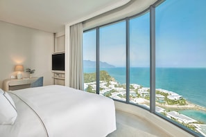 Une chambre d’hôtel avec un grand lit, une télévision, une table de chevet avec une lampe, et une vue sur la mer et les bâtiments.