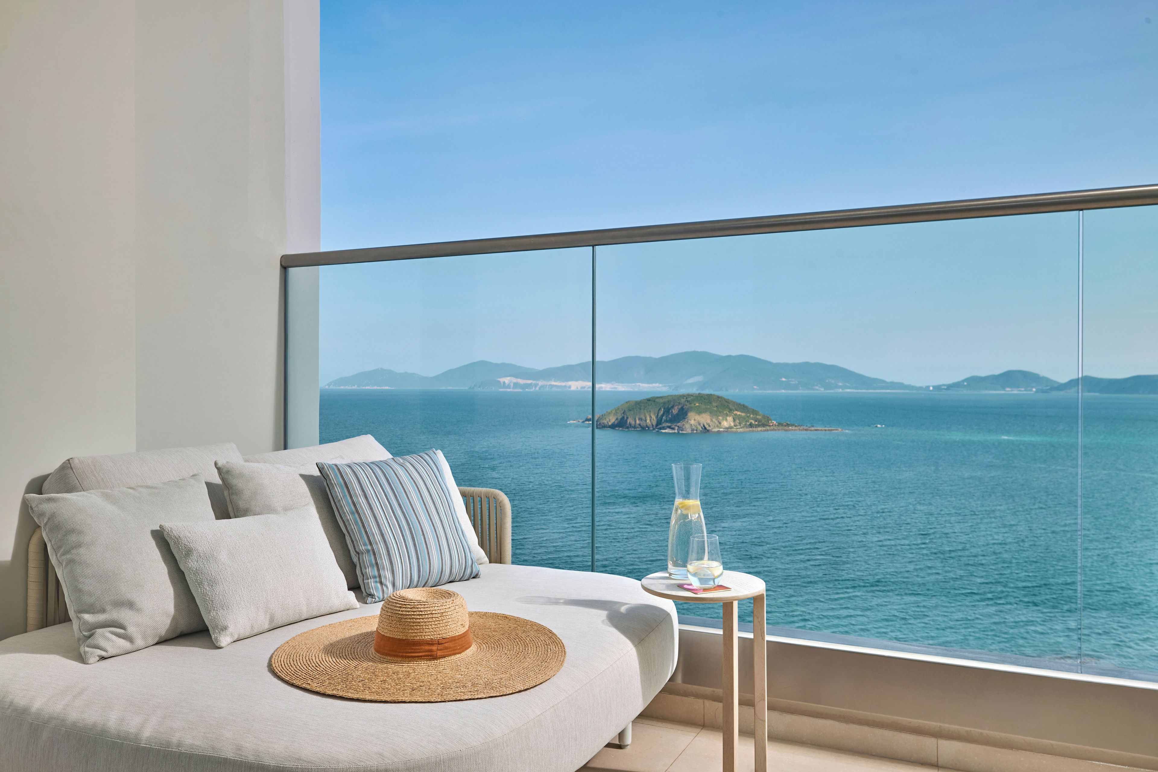A modern room with a large window overlooking a coastal view, a sofa with cushions, a small table with a drink, and a straw hat.