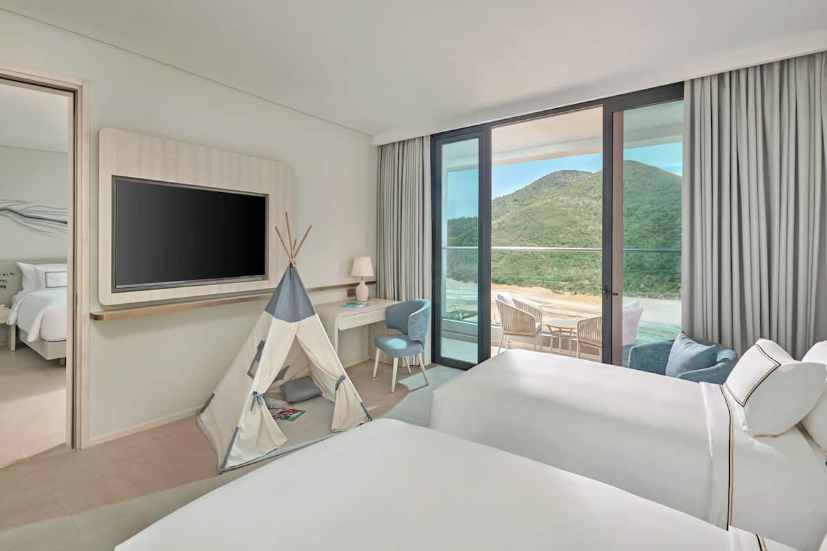 A hotel room with a bed, a desk, a chair, a TV, and a balcony with a view of the mountains.