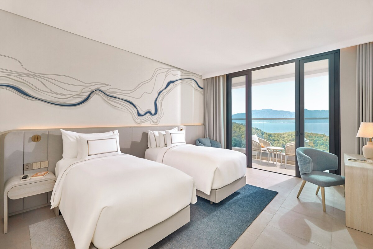 A hotel room with two beds, a desk, a chair, and a view of the outdoors.