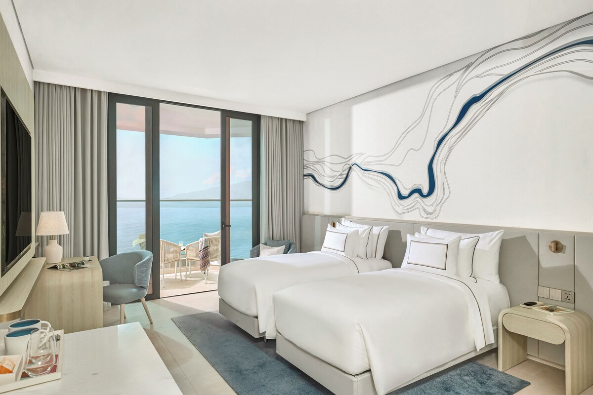 A hotel room with two beds, a desk, a chair, a TV, and a large window with a view of the sea.