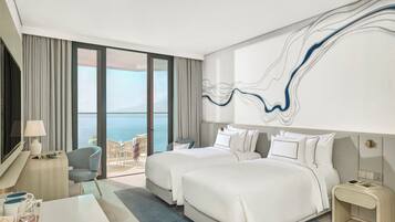 Une chambre d’hôtel avec deux lits, un bureau, une chaise, une télévision et une grande fenêtre donnant sur la mer.