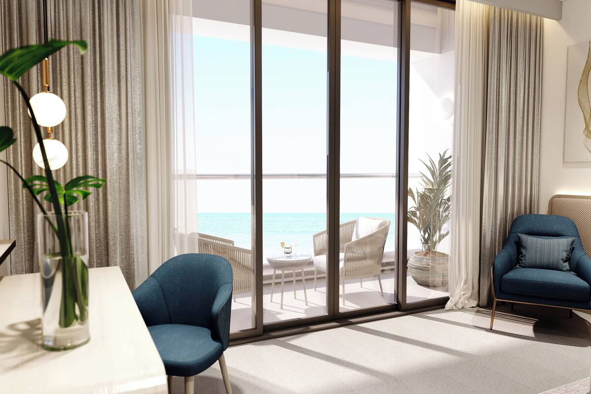 Un salon moderne doté d’une grande porte-fenêtre coulissante donnant sur un balcon avec vue sur la mer. On y trouve une table blanche ornée d’un vase contenant des plantes vertes, un fauteuil bleu et un canapé bleu avec un coussin.
