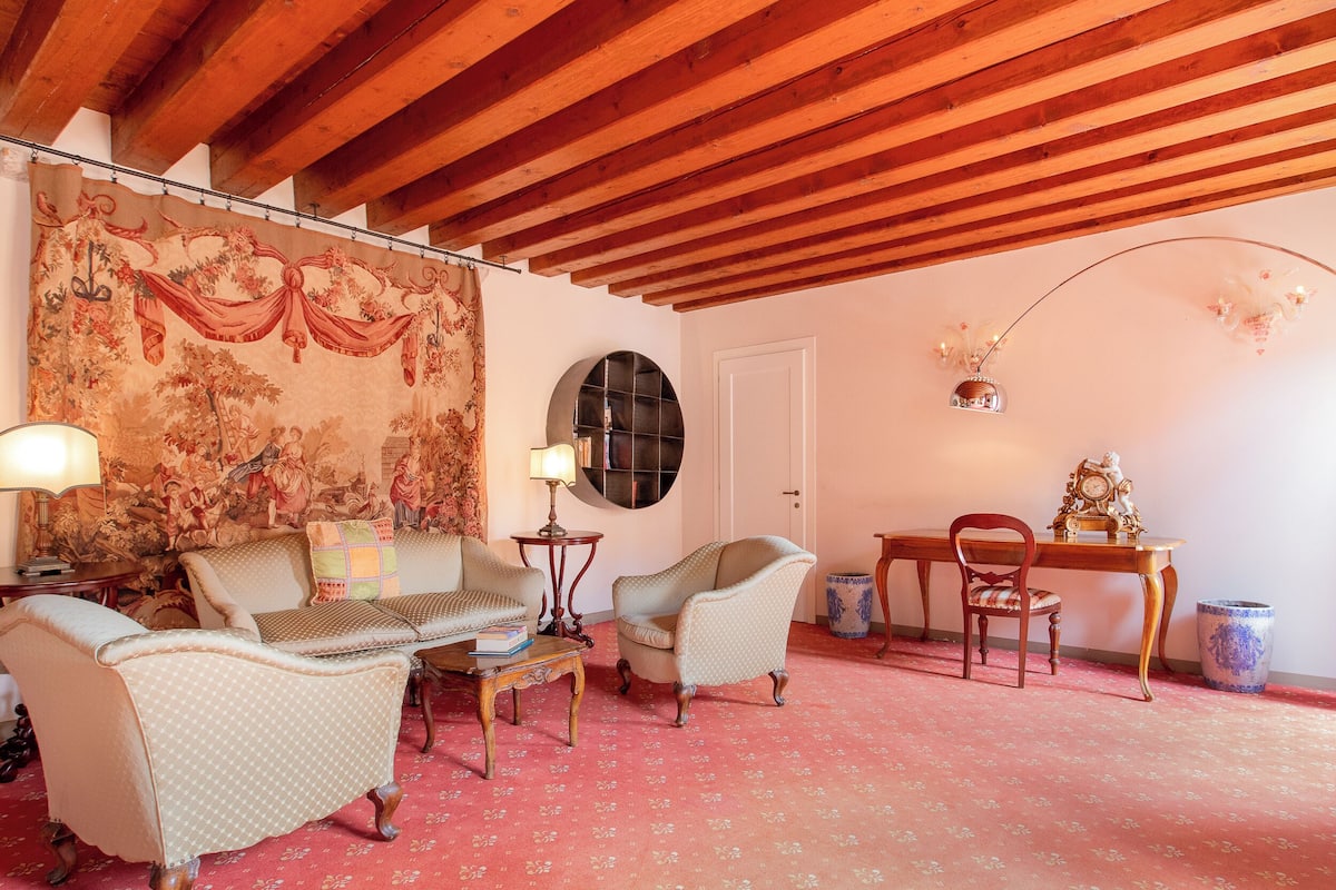 A room with a wooden ceiling, a tapestry, a round mirror, a table with a clock, and two chairs.