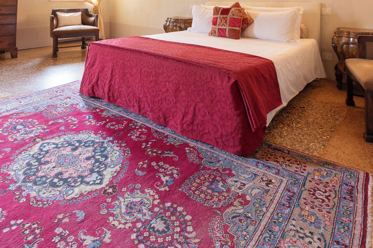 A bedroom with a wooden ceiling, a bed with a red bedspread, a patterned rug, and two wall-mounted lamps.