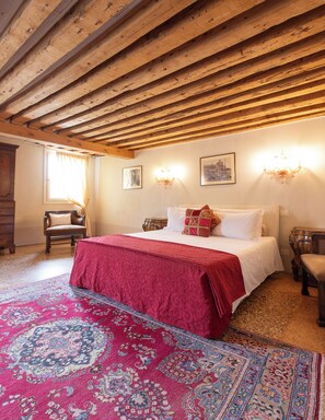 Une chambre avec un plafond en bois, un lit recouvert d’une courtepointe rouge, un tapis à motifs et deux lampes fixées au mur.