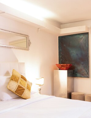 Une chambre d’hôtel avec un lit, un grand miroir, un tableau, un bol rouge, une lampe et deux poufs.