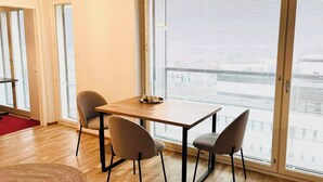 Ein Zimmer mit einem Holztisch, vier Stühlen, großen Fenstern mit Jalousien und Blick auf eine Stadtlandschaft.