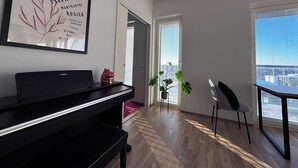 Ein modernes Wohnzimmer mit einem Klavier, einem gerahmten Kunstwerk, einer Pflanze und einem Essbereich mit Blick auf die Stadt.