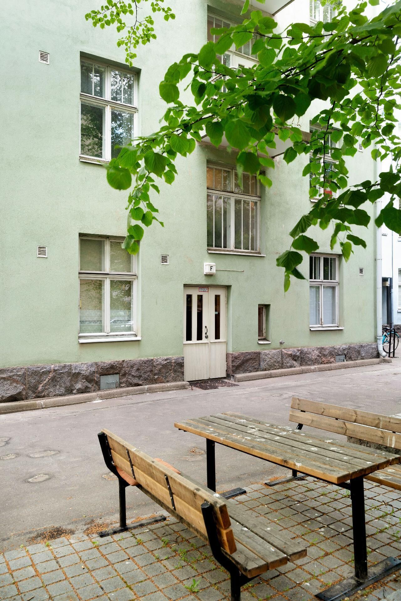 A building with a green facade, multiple windows, a door with a sign, and a paved area with two benches.