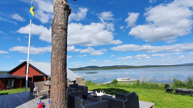 Una terraza de madera con mobiliario exterior, un asta con una bandera y una vista de un lago y montañas al fondo.