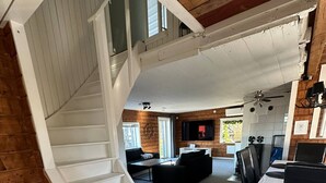 Un salón amplio con una escalera blanca que conduce a un nivel superior, un televisor de pantalla plana grande y un comedor con mesa y sillas.