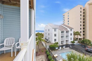 A balcony with white chairs, a pool area, and a view of a multi-story building with balconies.