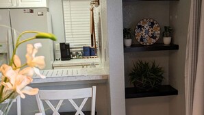 Una piccola cucina con mobili bianchi, una sedia bianca e un tavolo blu. Si vedono scaffali con oggetti decorativi e una pianta in vaso.
