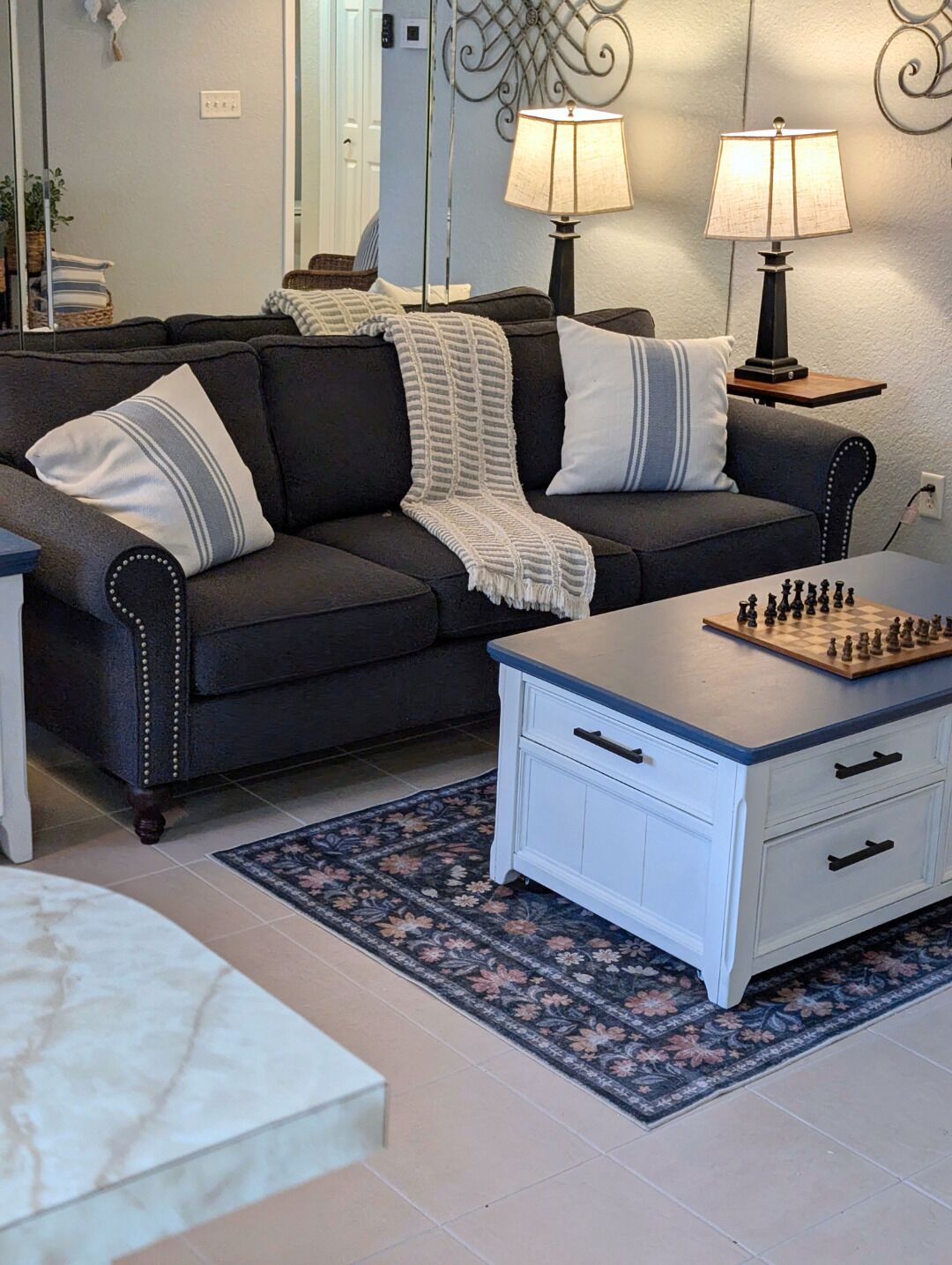 Ein Wohnzimmer mit einem dunkelgrauen Sofa, zwei Lampen, einem Schachbrett und einem floralen Teppich.