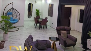 A modern living room with a sofa, a hanging chair, a dining table with chairs, and a potted plant. - Dwellux Apartment (Abuja)