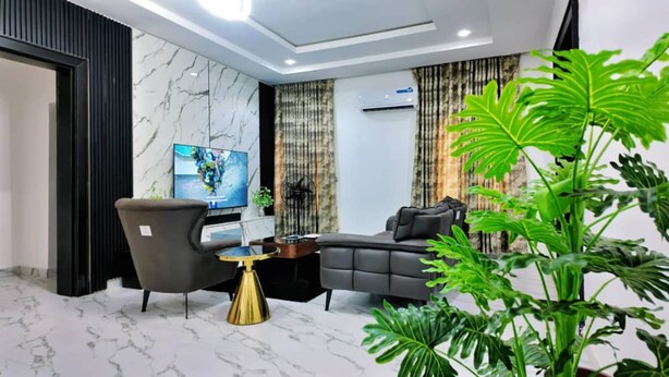 A modern living room with a marble floor, a grey sofa, a black armchair, a TV, and a large green plant. - Dwellux Apartment (Abuja)