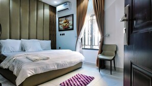 A hotel room with a large bed, a chair, a window with curtains, an air conditioning unit, and a painting on the wall. - Dwellux Apartment (Abuja)