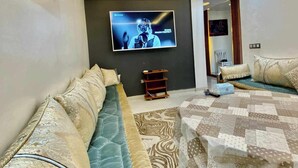 A modern living room with a sofa, a bed, a TV mounted on the wall, and a small wooden shelf. - Apartment agadir bay flat luxe (Agadir)