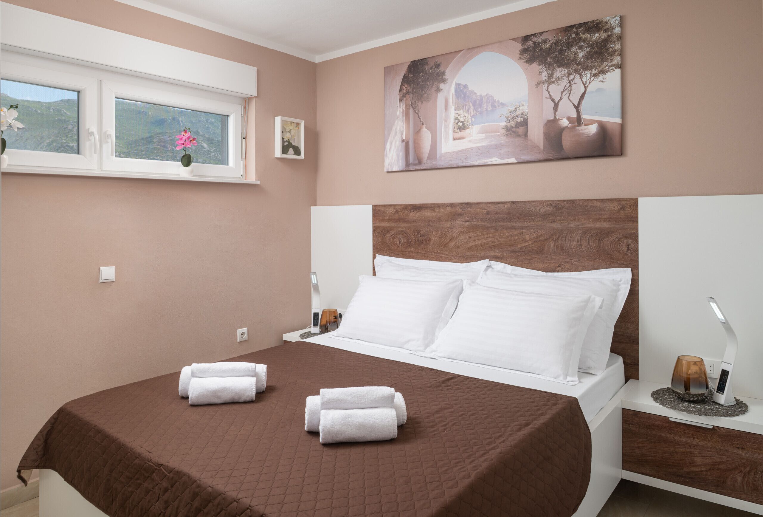 Une chambre à coucher avec un lit, des oreillers, une table de chevet, une lampe, une fenêtre décorée de fleurs et un tableau au mur.