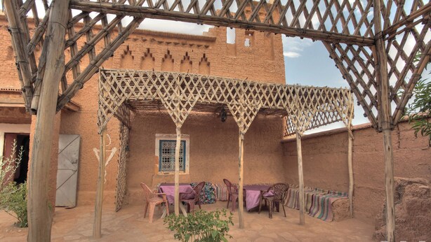 An outdoor seating area with a wooden lattice structure, a table with chairs, and a small plant in the foreground. - Maison d'hôtes tifaoute ait Ben haddou  (Ait Benhaddou)