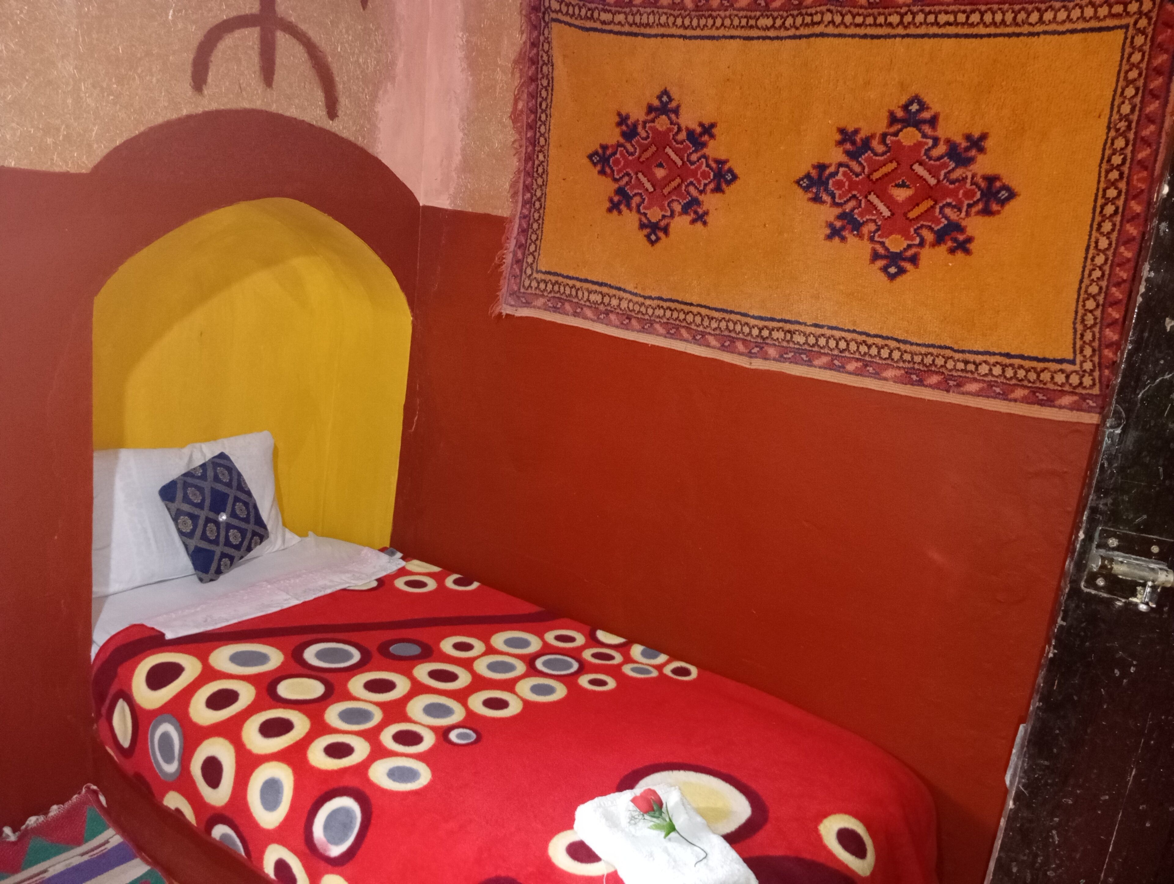Een kamer met een rood en geel sprei, een gestreept kussen, en een decoratieve wandversiering.