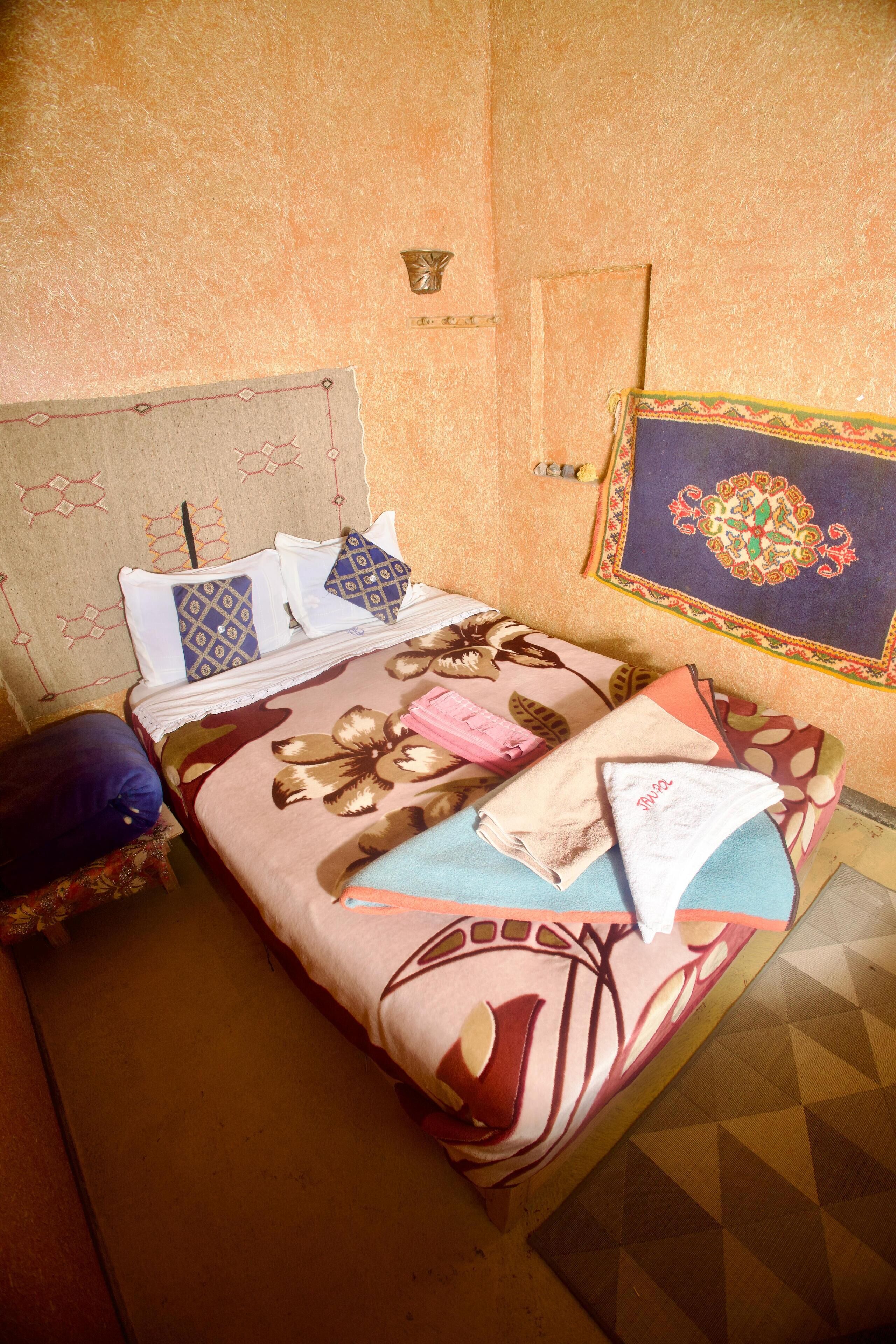 A neatly made bed with patterned bedding and pillows, a small bedside table with a lamp, and a decorative wall hanging.
