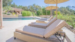Un espace aménagé au bord de la piscine, avec des chaises longues et des parasols, une piscine, et un bâtiment doté d’un toit recouvert de tuiles.