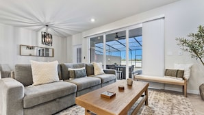 A modern living room with a grey sectional sofa, a wooden coffee table, and a bench. There is a large mirror on the wall, a ceiling fan, and a view of the outdoors through large windows. - 16148 Palm Frond Avenue (Clermont)