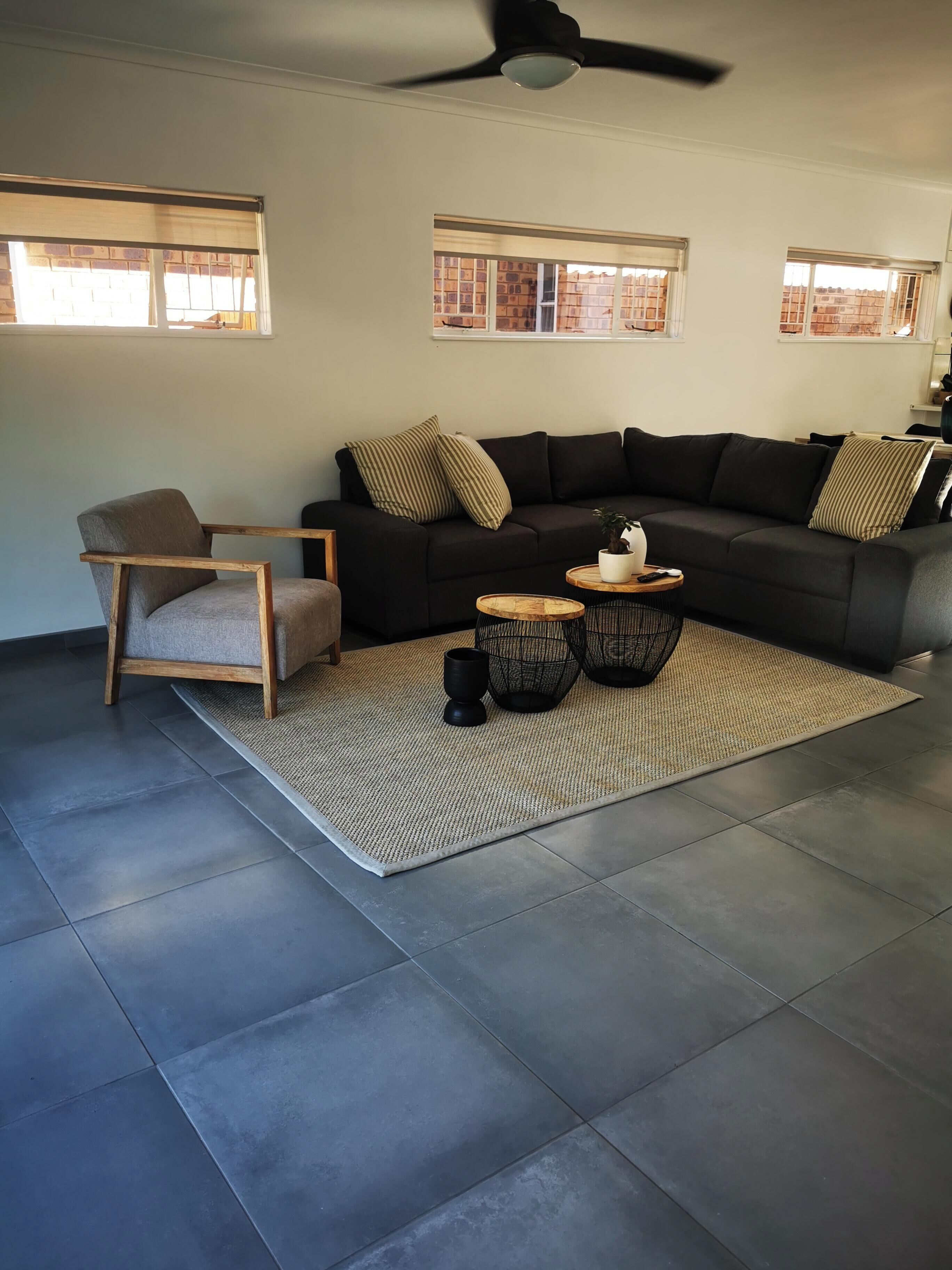 A modern living room with a dark-colored sofa, a wooden chair, a woven rug, and a ceiling fan.