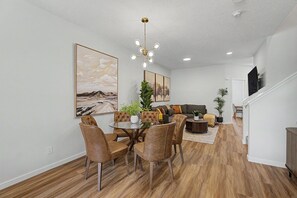 Ein Essbereich mit einem runden Tisch, Stühlen und einer Chandelüster. Im Hintergrund ist ein Wohnzimmer mit einer Couch und einem Fernseher zu sehen.