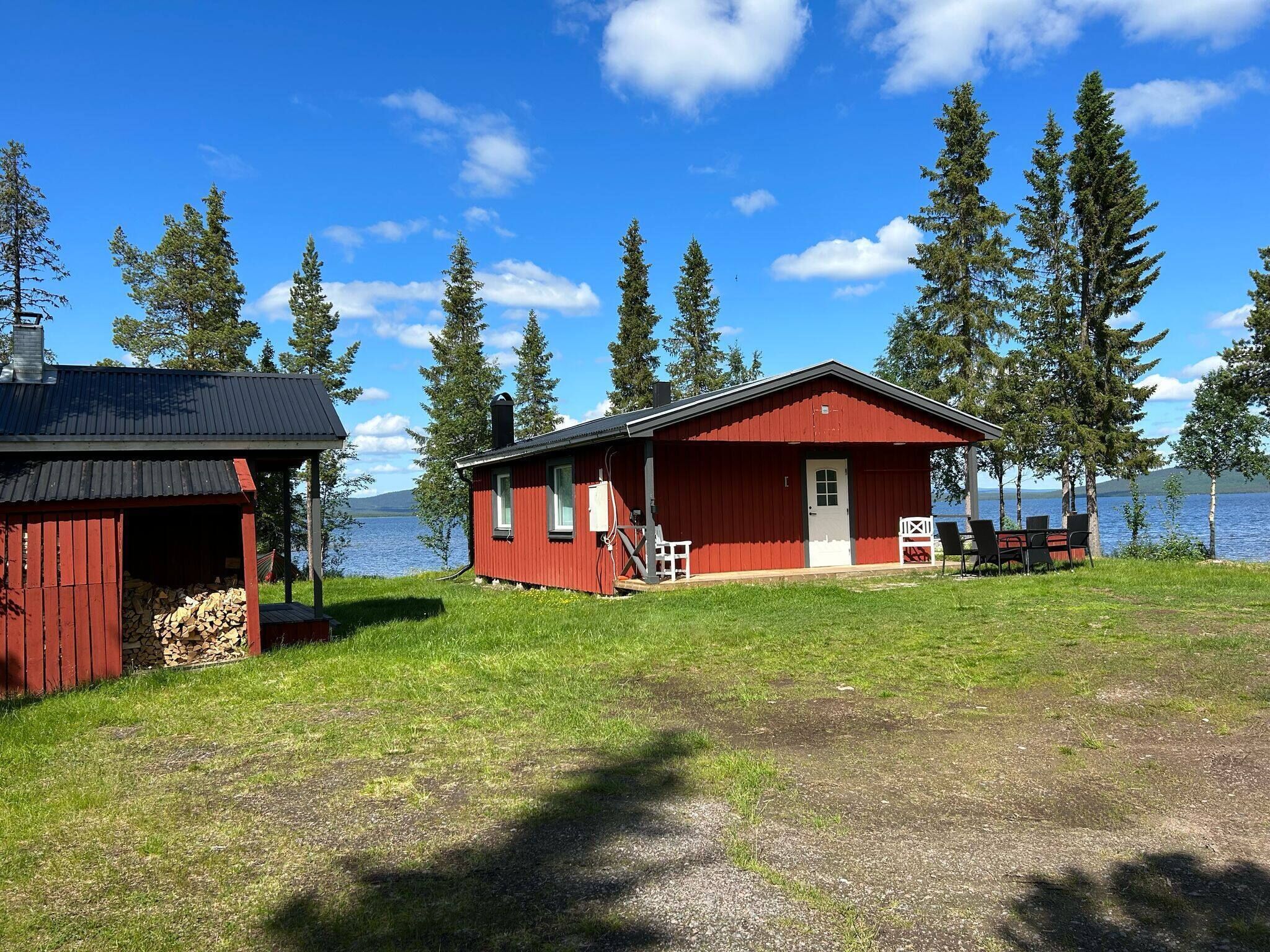 A red cabin with a porch and a small deck, surrounded by trees and a lake.