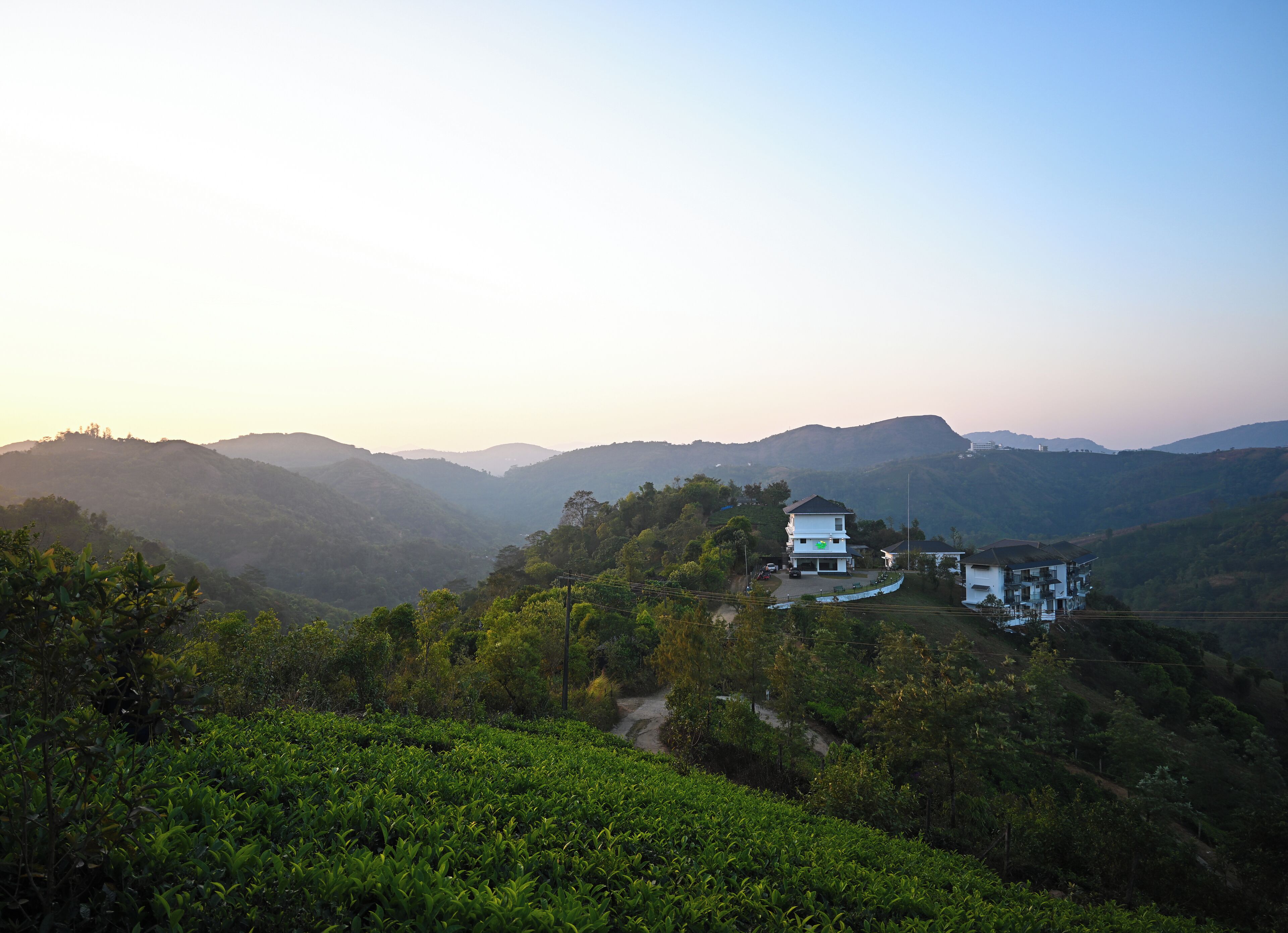 A two-story building with a flat roof and a balcony, surrounded by lush greenery and hills in the background.