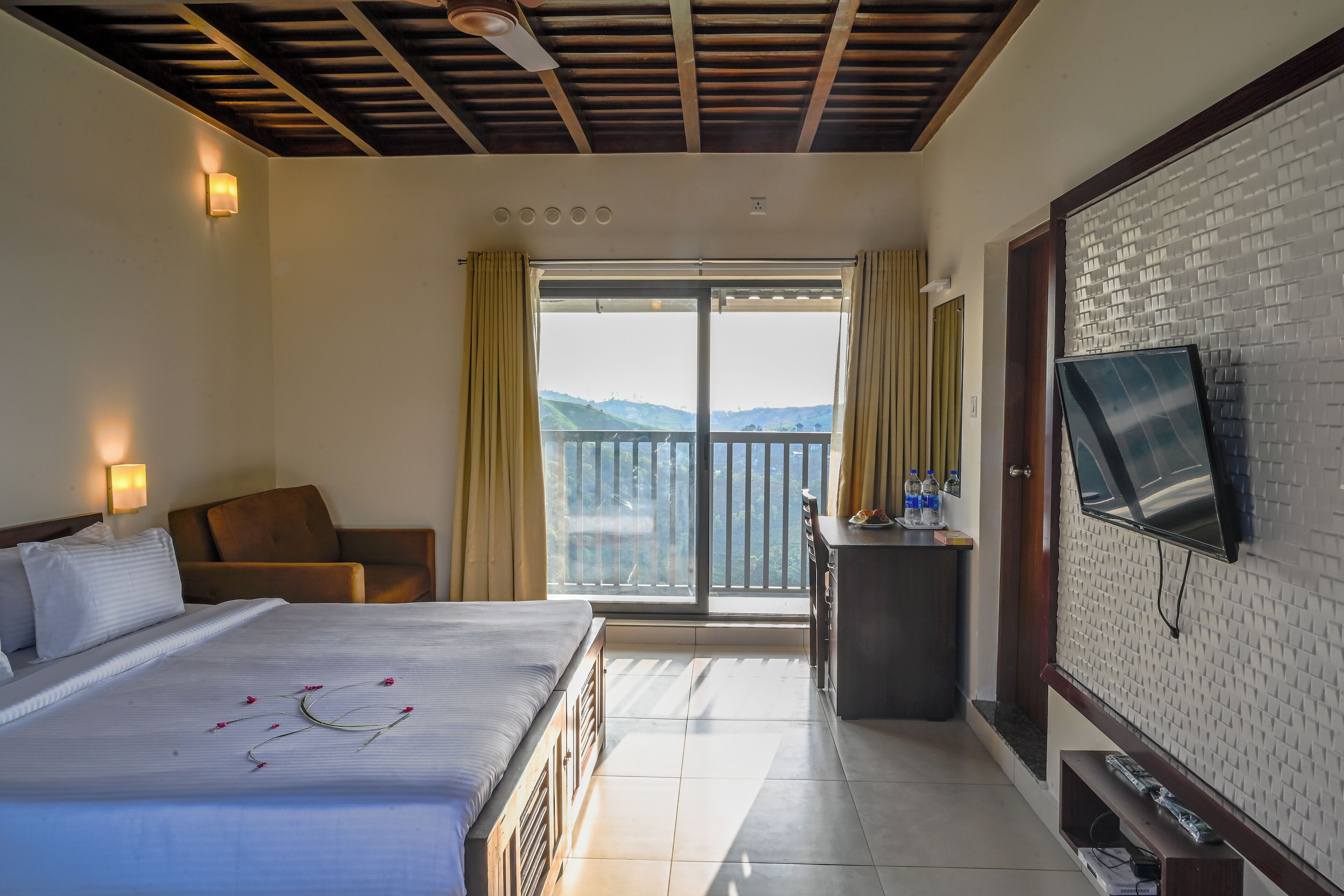A hotel room with a bed, a TV mounted on the wall, a balcony with a view, and a desk with a mirror and toiletries.