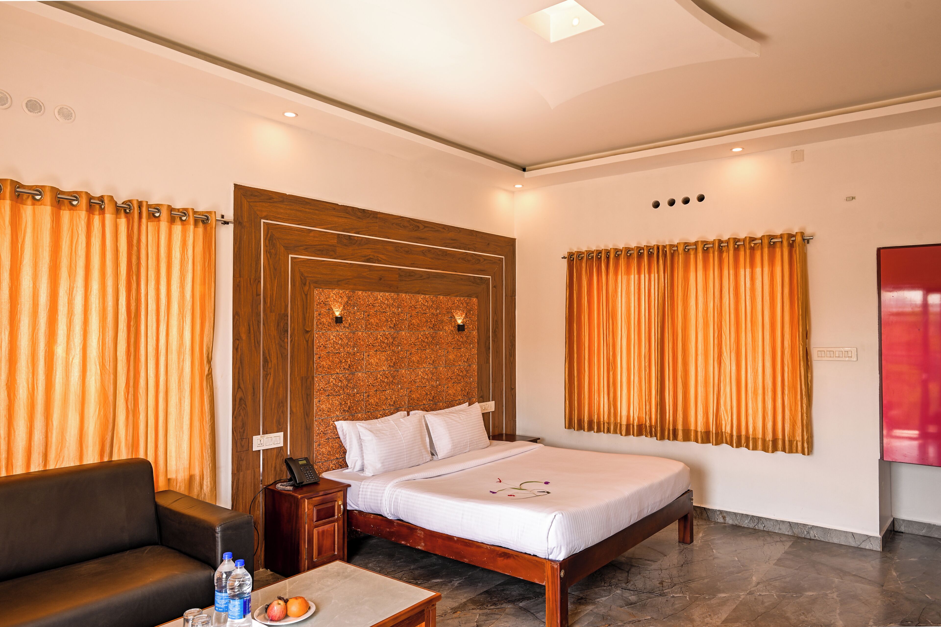 A modern hotel room with a wooden headboard, a bed with white linens, a leather sofa, a small table with a water bottle and fruit, and a red artwork on the wall.