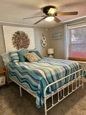 A bedroom with a bed, a ceiling fan, a wall-mounted decoration, a bedside table with a lamp, and a window with blinds.