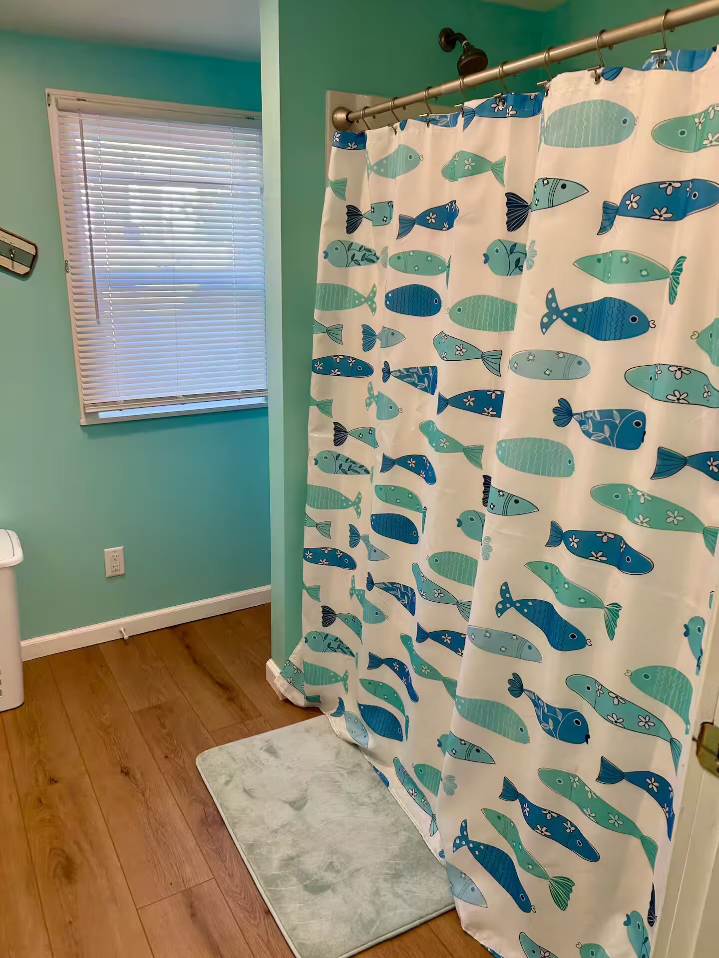 A bathroom with teal walls, a shower curtain with fish patterns, a wooden floor, and a light-colored rug.