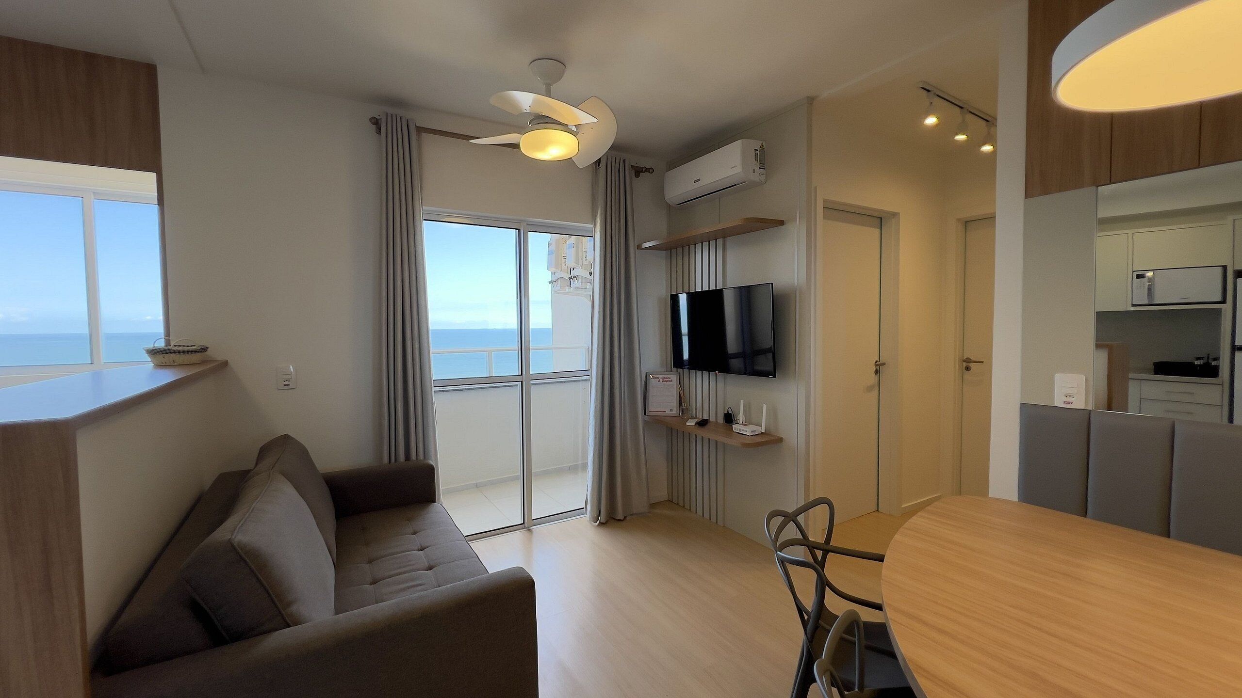 A modern living room with a sofa, a dining table, a TV mounted on the wall, and a view of the sea through large windows.