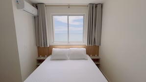 A hotel room with a bed, a window, curtains, a wooden headboard, and a wall-mounted air conditioner. - Solarium Beach Club -Apartamento T1.1105 (Itapoá)