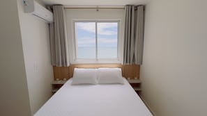A hotel room with a bed, a window, curtains, a wooden headboard, and a wall-mounted air conditioner.