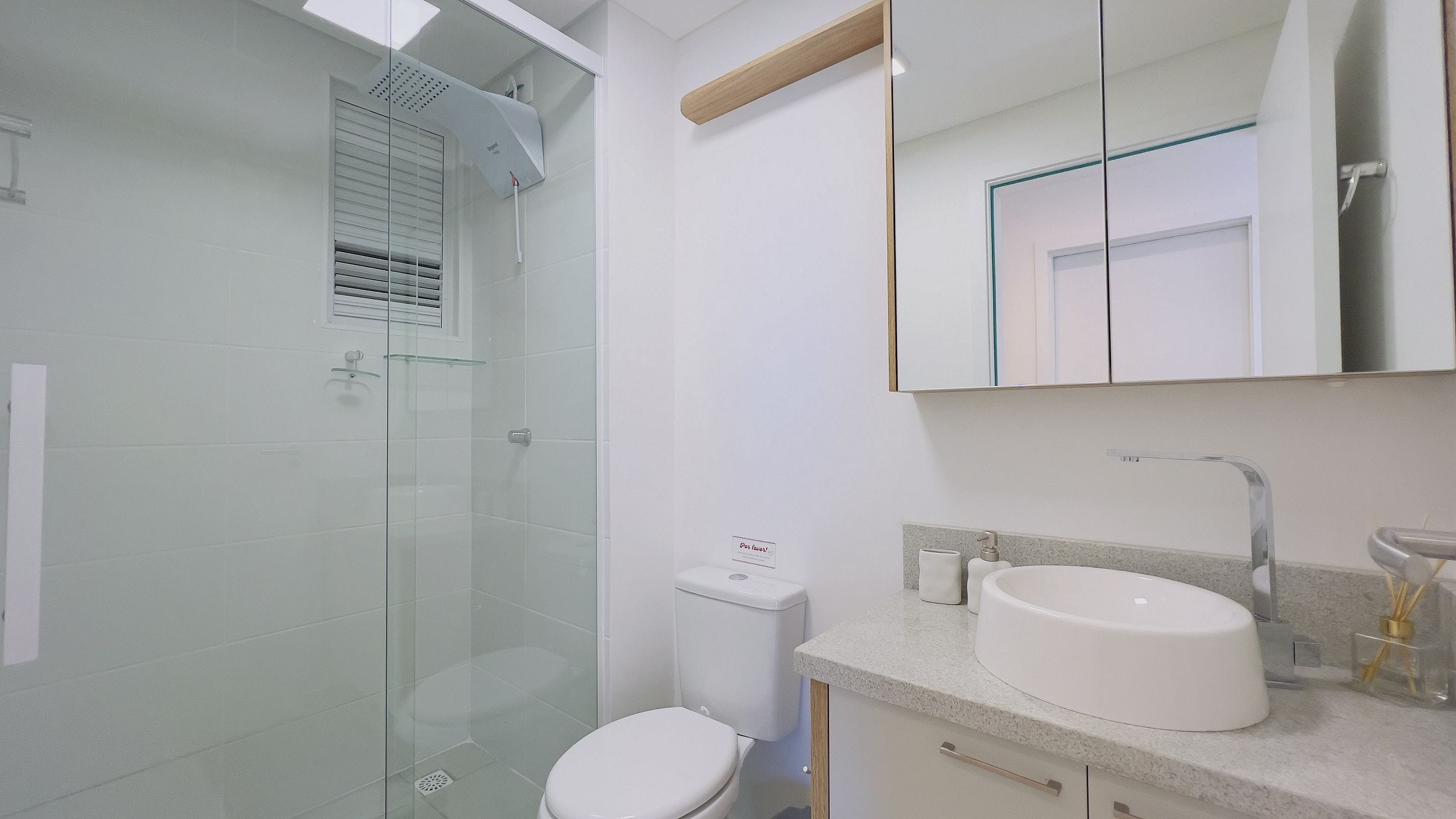 Un baño moderno con cabina de ducha de vidrio, inodoro, lavabo con espejo y una ventana pequeña con persianas.