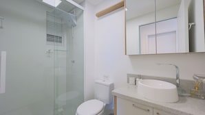 Ett modernt badrum med en duschkabin i glas, en toalett, ett handfat med en spegel ovanför och ett litet fönster med persienner.