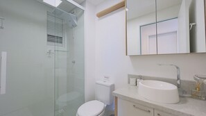 A modern bathroom with a glass shower enclosure, a toilet, a sink with a mirror above it, and a small window with blinds. - Solarium Beach Club -Apartamento T1.1105 (Itapoá)